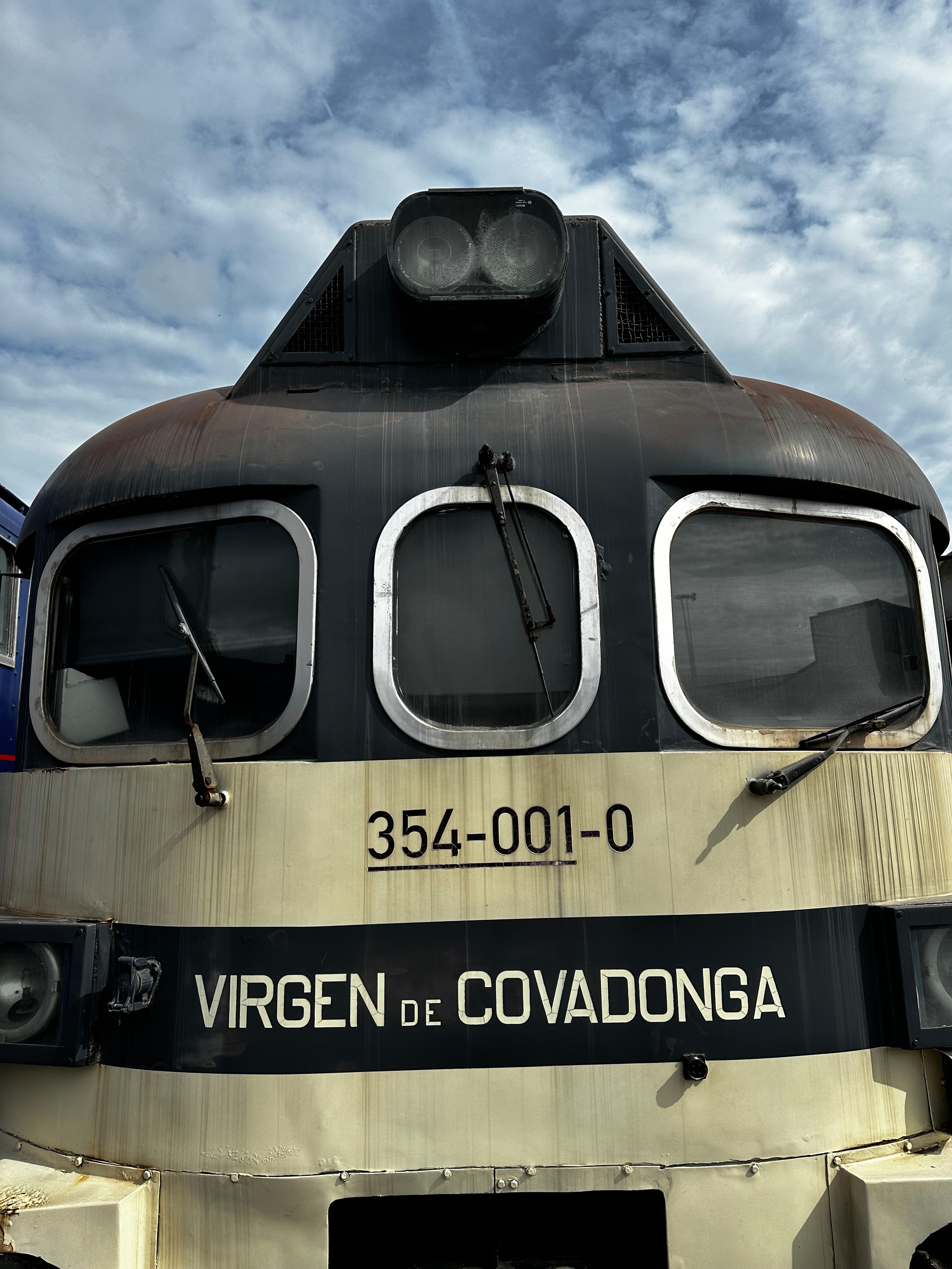 A close-up of the front of a black and beige train labeled Virgen de Covadonga with a cloudy sky in the background.