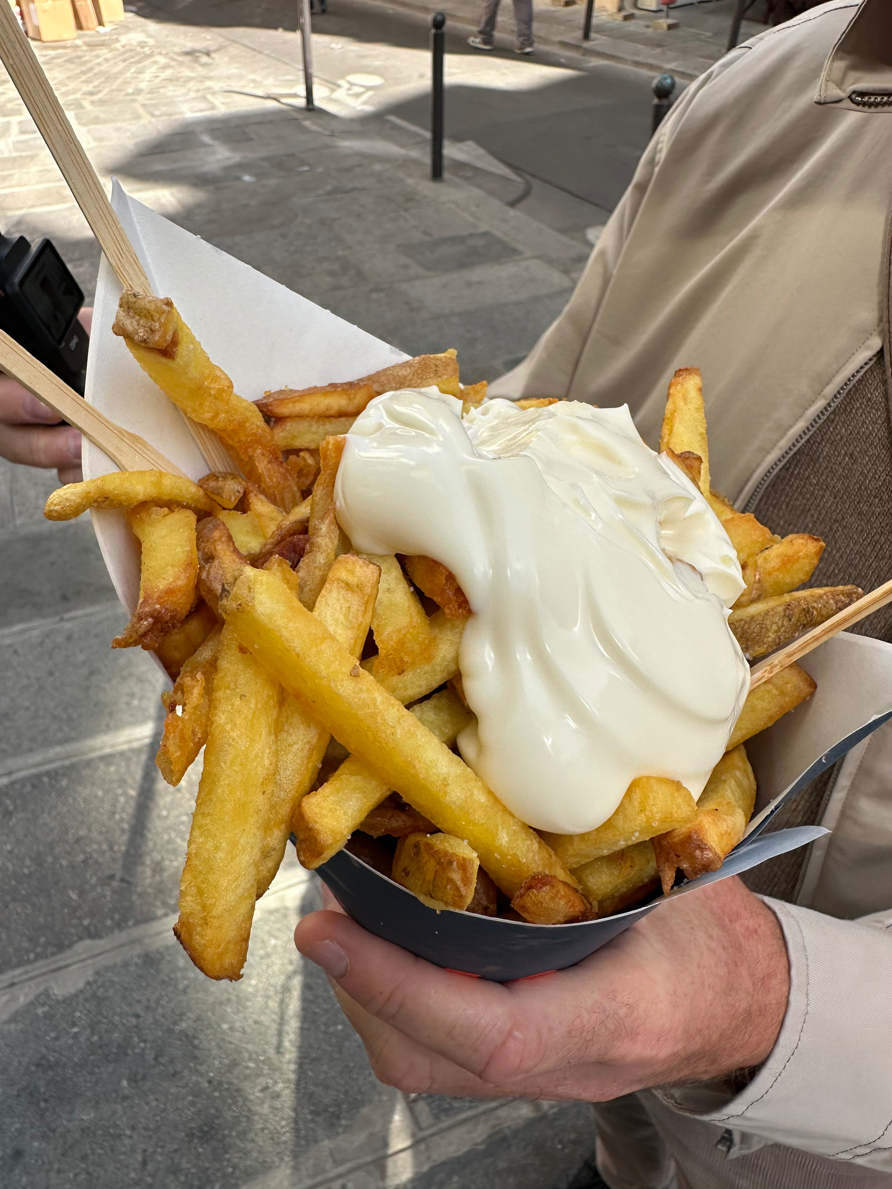 A person is holding a serving of thick-cut fries generously topped with a large dollop of creamy mayonnaise.
