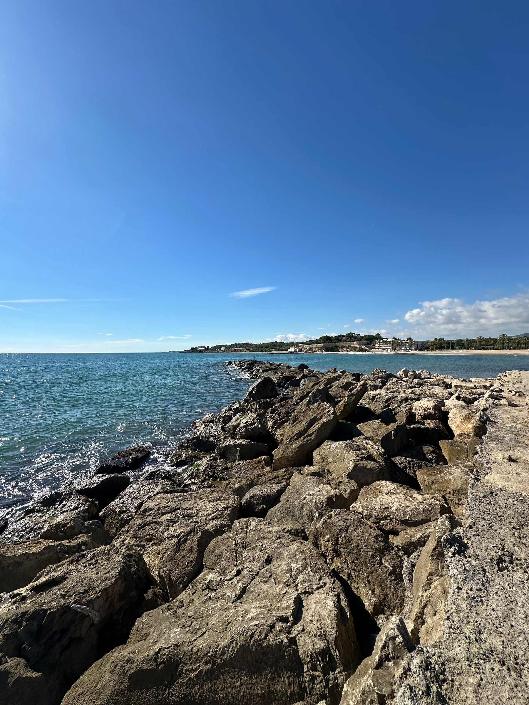 A rocky breakwater extends into a calm sea under a clear blue sky with distant shorelines visible on the horizon.