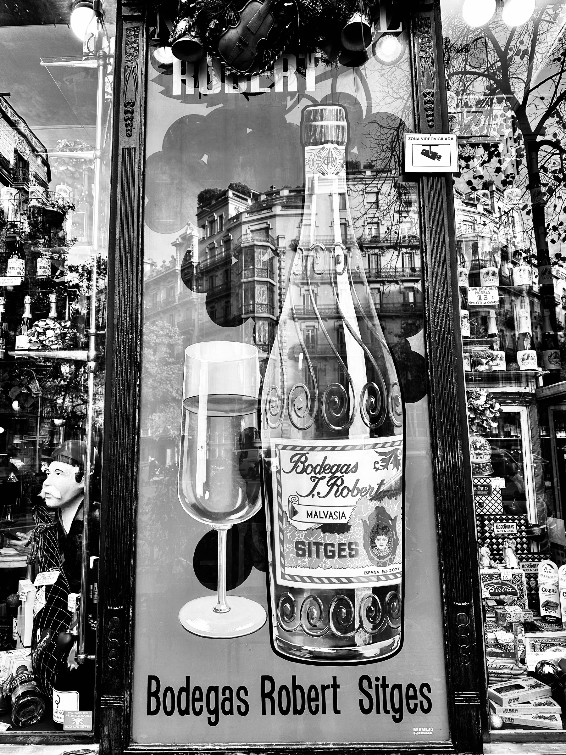 A black and white window display features a large advertisement for Bodegas Robert Sitges with a bottle, a glass, and reflective architecture.