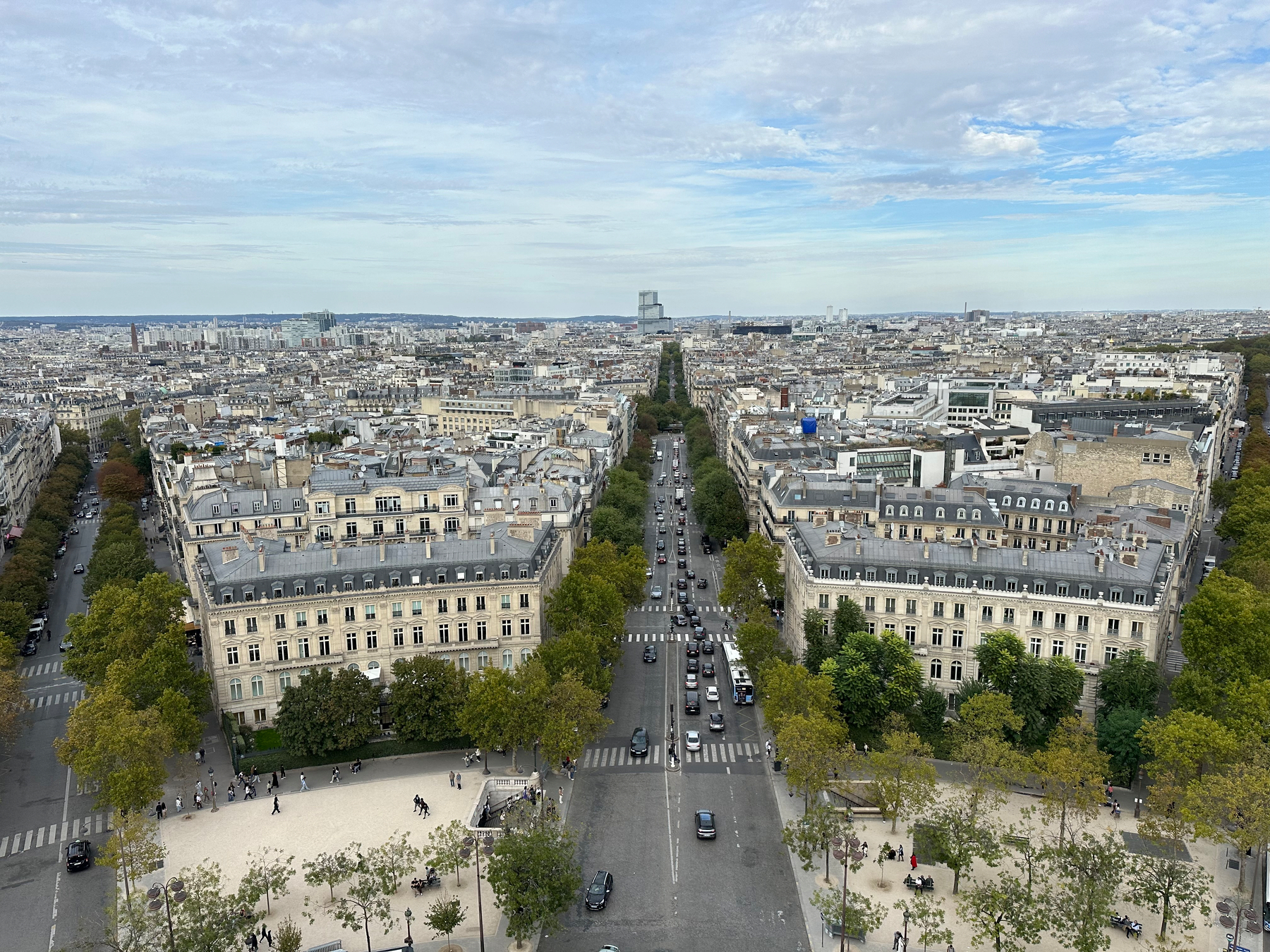 A wide aerial view of a cityscape with tree-lined streets, historic buildings, and a busy road stretching into the distance under a partly cloudy sky.