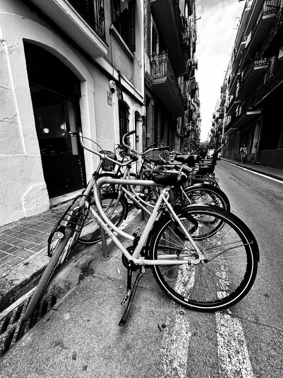Several bicycles are parked and locked to a rack along a narrow street lined with buildings, captured in black and white.